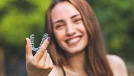 Young woman with purple nails smiling holding clear aligner with two fingers