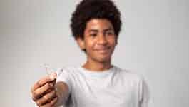 Teenager in grey shirt holding out clear aligner in front of gray faded background